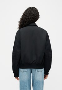 Άτομο με μακριά σγουρά μαλλιά, ντυμένο με μαύρο bomber μπουφάν και ανοιχτόχρωμα μπλε τζιν, στέκεται γυρισμένο με την πλάτη προς το απλό φόντο.