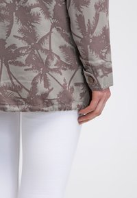 Långärmad skjorta med palmtryck i dämpad beige och brun med knappränder och samlad nederkant. Matchad med figurnära vita byxor.