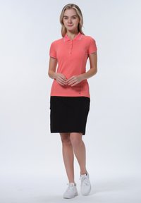 Růžová polo košile s límečkem a knoflíkovou légou, v kombinaci s černou sukní ke kolenům. Model má na sobě bílé tenisky. Hladká textura.