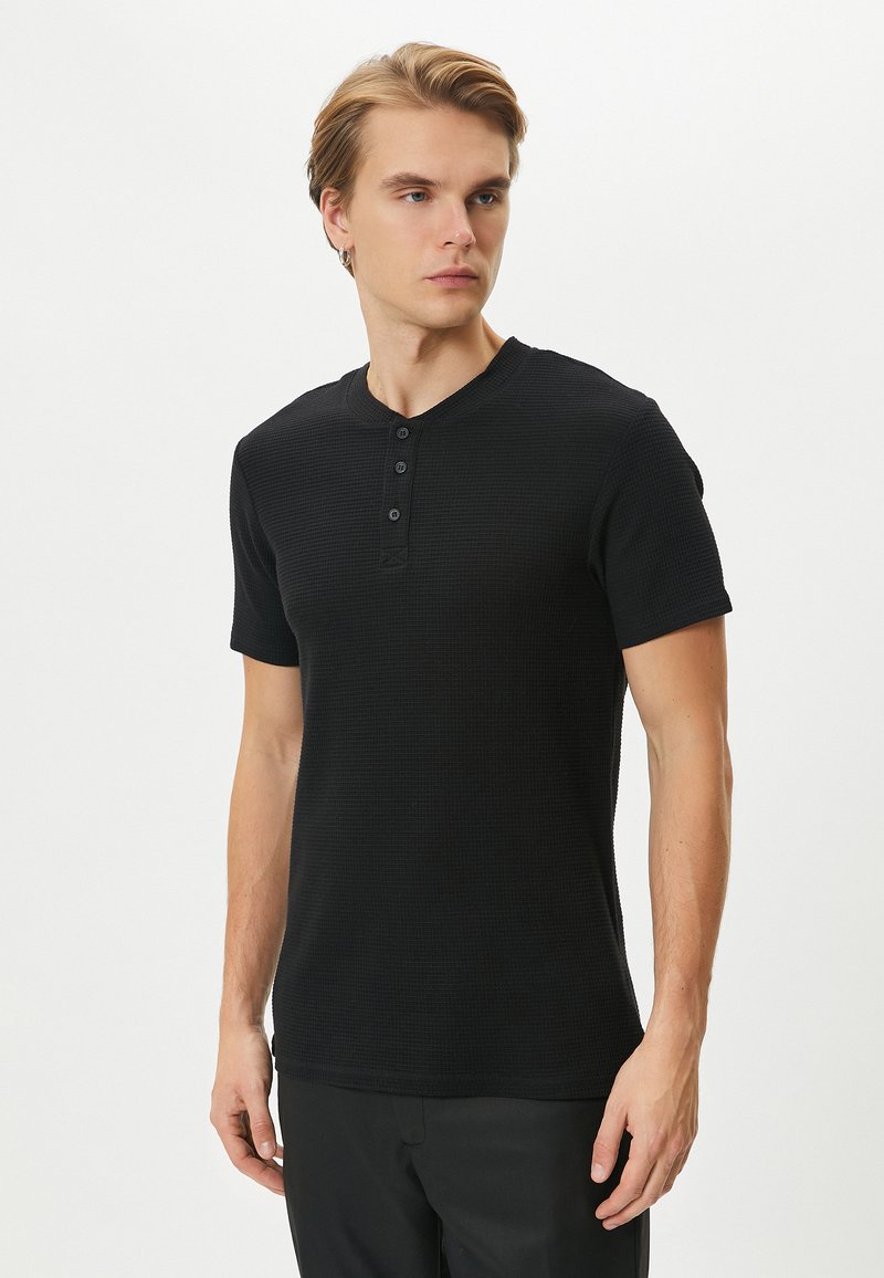 Camisa henley de manga corta negra con una tela texturizada, que presenta una tapeta de tres botones y un sutil diseño de canalé.