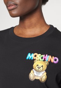 Felpa nera con un logo colorato "MOSCHINO" e una grafica di un orsetto peluche, che presenta un'etichetta con la scritta "QUESTO NON È UN GIOCATTolo MOSCHINO."