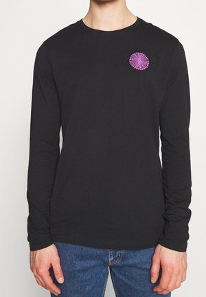 Chemise noire à manches longues avec un petit motif rond rose sur le côté gauche de la poitrine, portée par une personne portant un jean bleu.