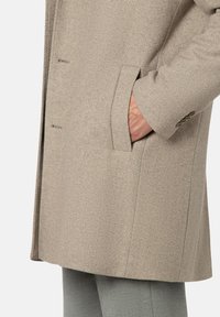 Beiger Mantel aus strukturiertem Stoff mit seitlicher Tasche, tailliertem Schnitt und dezenten Steppnähten. Silhouette reicht bis zur Mitte des Oberschenkels.