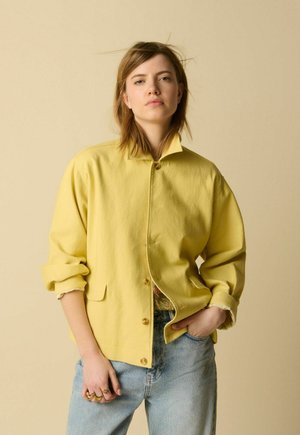 Jeune femme aux cheveux châtain clair portant une veste jaune boutonnée et un jean bleu clair, debout avec une main dans la poche.