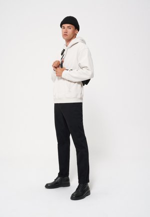 Witte hoodie, zwarte broek en zwarte veterlaarzen. Effen kleur, relaxte pasvorm en grote voorzak. Zwart beanie-accessoire.