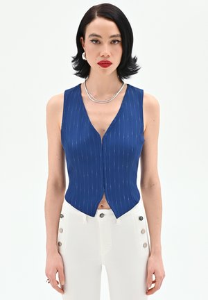 Donna con capelli corti neri che indossa un gilet blu a righe sottili senza maniche e pantaloni bianchi a vita alta con bottoni laterali, su sfondo bianco.