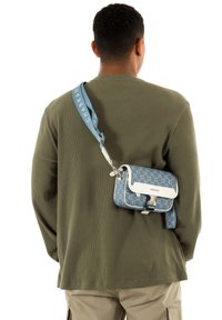 Blauwe crossbodytas met een patroon en witte accenten, verstelbare schouderband en metalen hardware, gedragen over een olijfgroene longsleeve.