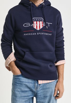 Mężczyzna noszący granatową bluzę z kapturem GANT z emblematem i napisem "American Sportswear" na jasnoróżowej koszuli w paski i niebieskich dżinsach.