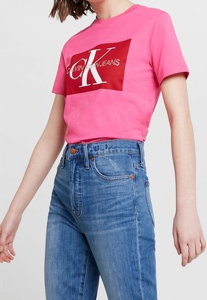 Rosa Baumwoll-T-Shirt mit rotem Grafikfeld, auf dem "CALVIN KLEIN JEANS" und ein weißer Buchstabe "K" abgebildet sind. Kombiniert mit hoch taillierten blauen Jeans.
