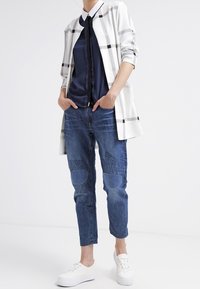 Cardigan largo de cuadros en blanco y gris, camiseta azul oscura, jeans de mezclilla con parches estampados y zapatillas blancas. Conjunto casual con texturas variadas.