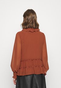 Rostfärgad blus med långa, puffiga ärmar, volangkrage och peplum midja. Tillverkad av lätt tyg med en mjuk textur.