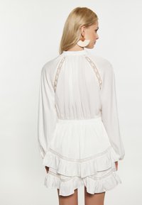 Witte jurk met lange mouwen, kanten details op de rug en taille, gelaagde ruffle zoom, gemaakt van een lichte stof met een gladde textuur.