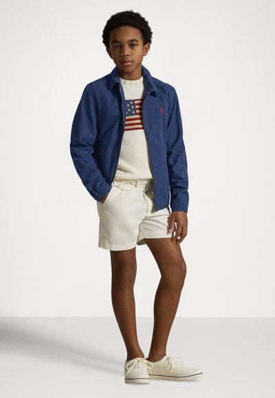 Tamsiai mėlynas džinsinis švarkelis ant kreminės spalvos megztinio su Amerikos vėliavos dizainu, kreminės spalvos šortai ir šviesios spalvos batai. Paprasta ir kasdienė apranga.