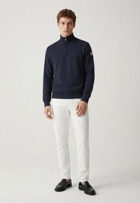 Marineblauer Sweatshirt mit Reißverschluss aus weichem Stoff, mit Stehkragen, gerippten Bündchen und einem Logo auf dem Ärmel, kombiniert mit weißen Hosen und schwarzen Slippern.