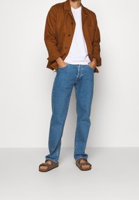 Man som bär en brun jacka, vit t-shirt, blå jeans och bruna sandaler står med händerna i fickorna mot en neutral bakgrund.