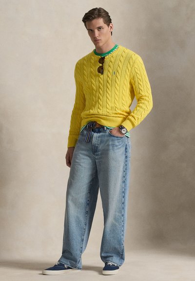 Rumeno pulover z vzorcem pletenja čez zeleno srajco, kombiniran z ohlapnimi svetlo modrimi kavbojkami in modrimi supergami. Vključene so tudi modne sončna očala in ura.