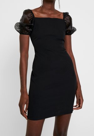 Kvinde iført en tætsiddende sort kjole med gennemsigtige pufærmer og firkantet halsudskæring, accessorized med lange hængende øreringe.