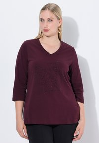 Frau mit glattem, blonden Haar, die ein burgunderfarbenes V-Ausschnitt-Shirt mit Dreiviertelärmeln und schwarze Hosen trägt, im Stehen vor einer weißen Wand.