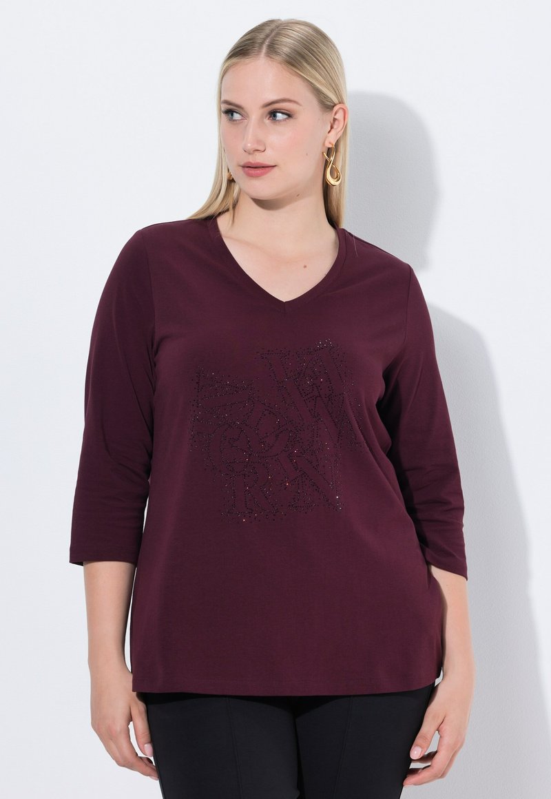 Frau mit glattem, blonden Haar, die ein burgunderfarbenes V-Ausschnitt-Shirt mit Dreiviertelärmeln und schwarze Hosen trägt, im Stehen vor einer weißen Wand.