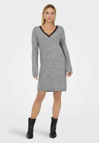 Vestido de punto gris con escote en V y detalles de encaje negro. Mangas largas y silueta recta. Llevado con botines negros.