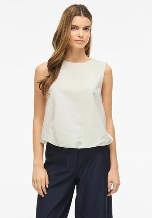 Ung kvinde med langt brunt hår iført en ærmeløs off-white top og mørke, vide bukser, stående mod en ensfarvet baggrund.