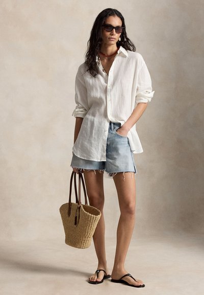 Nő túlságosan nagy fehér ingben, farmer rövidnadrágban, fekete szandálban, napszemüvegben, piros nyaklánccal, kezében fonott szalmakosárral, semleges háttér előtt.