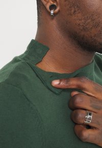 Grön stickad tröja med en unik kragdesign som har en vikbar kant. Närbild visar en hand som pekar på kragsdetaile.