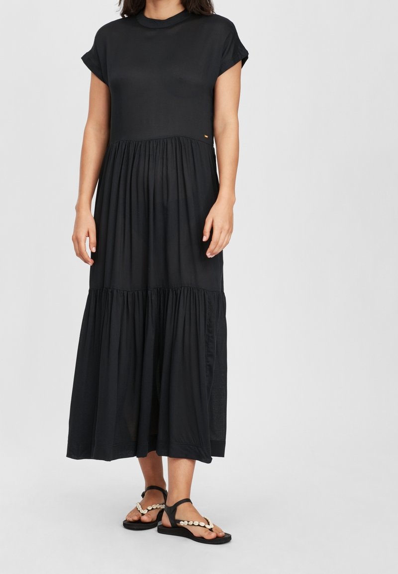 Femme portant une robe midi noire à manches courtes avec une jupe à volants et des sandales noires décorées de coquillages blancs sur un fond uni.