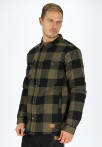 Long-sleeve, button-up shirt in black and olive green checkered pattern. Features two chest pockets and a fabric label at the bottom.