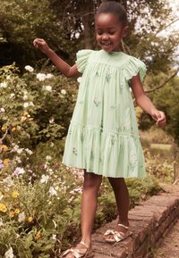 Vihreä mekko, jossa on röyhelöhiha, kukkakirjonta ja kerroksellinen helma. Lapsella on päällään kultaiset sandaalit ja hän seisoo kukkien keskellä.