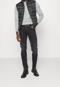 Chaleco de plumas negro sobre un suéter gris claro, combinado con jeans oscuros y botas de cuero negras hasta el tobillo. Presenta detalles de cremallera y cinturón.