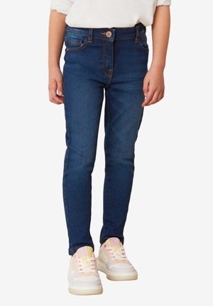 Enfant portant un jean skinny bleu foncé et des baskets aux couleurs pastel, debout avec les bras détendus le long du corps sur un fond blanc.