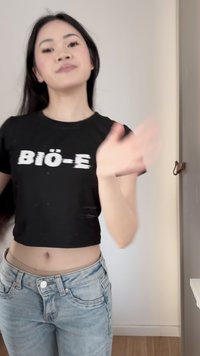 Junge Frau mit langen dunklen Haaren, die ein schwarzes kurzes Oberteil und blaue Jeans trägt und in einer neutralen Innenumgebung die Hand winkt.