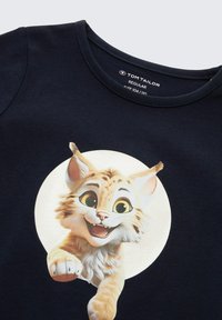 Natürlichem Baumwoll-T-Shirt in Marineblau mit einem strukturierten, glänzenden Grafikdesign eines lächelnden Katers mit großen Augen, das sich in einem kreisförmigen, hellen Bereich befindet.