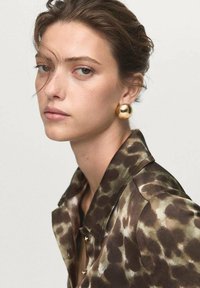 Hnědá košile s leopardím potiskem, hladká textura; zlaté kulaté náušnice; přirozený make-up; neutrální pozadí.