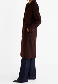 Hnědý vlněný kabát s límcem, pěti knoflíky, bočními kapsami a klasickým designem, v kombinaci s džíny volného střihu a tmavými botami.