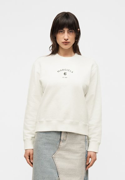 Femme aux cheveux foncés mi-longs porte un sweat-shirt blanc avec l’inscription « MARGIELA 6 EST 1997 » et une jupe en jean bicolore.