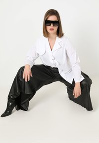 Schwarze Kunstleder-Hosen mit weitem Bein, weißes Hemd mit großen Kragen und silbernem Ringdetail, kombiniert mit übergroßen schwarzen Sonnenbrillen.