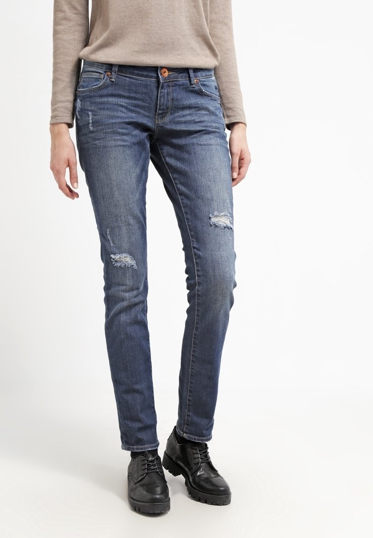 Personne portant un jean skinny bleu déchiré, un haut beige à manches longues et des chaussures noires à lacets, debout devant un fond blanc.