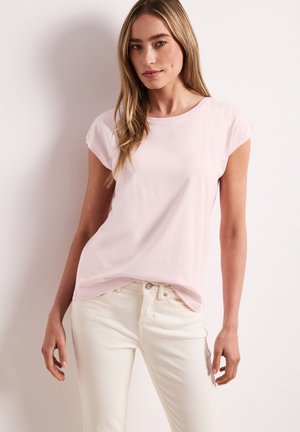 Jeune femme aux longs cheveux blonds portant un t-shirt rose pâle à manches courtes et un jean blanc, debout devant un fond clair.