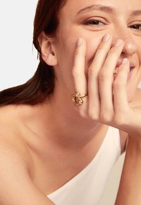 Anillo de oro con un diseño único y abstracto que presenta acentos redondeados y suaves. El modelo lo lleva en la mano izquierda, cubriendo parcialmente su rostro.