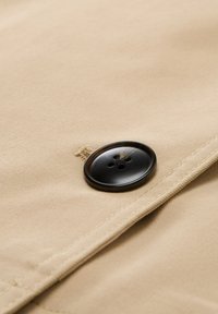 Beiger Stoff mit glatter Textur zeigt einen großen, glänzenden schwarzen Knopf mit vier Löchern. Die Nähte sind dezent und gut verarbeitet.