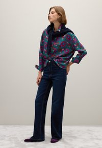 Frau steht da, trägt dunkelblaue Jeans, lila Schuhe, ein bunt gemustertes Hemd mit Blumenmuster und einen marineblauen Pullover über den Schultern.