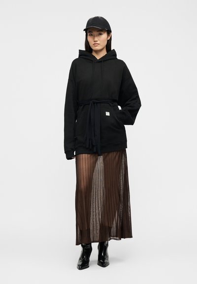 Γυναίκα που φοράει μαύρο φόρεμα-φούτερ με δεμένα γάντια στη μέση, μια διαφανή μακριά φούστα, μαύρες μυτερές μπότες και μαύρο καπέλο μπέιζμπολ.