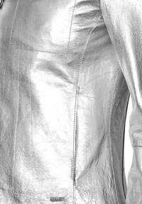 Srebrna skórzana kurtka o gładkiej, błyszczącej fakturze z widocznymi przeszyciami i firmową metką na dolnym brzegu.