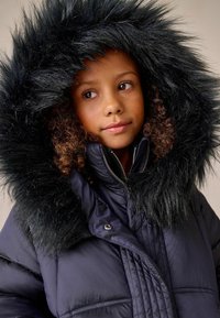 Sötétkék puff coat, nagy fekete műszőrme kapucnival. Cipzáros és patentos záródás, steppelt textúra és sötét gombok jellemzik.