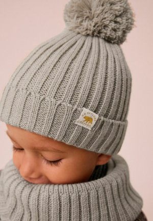 Beanie di maglia grigio chiaro con una texture a coste e un morbido pon-pon sopra. Presenta una patch con un orso e il testo "VAI A ESPLORARE ALL'ESTERNO".