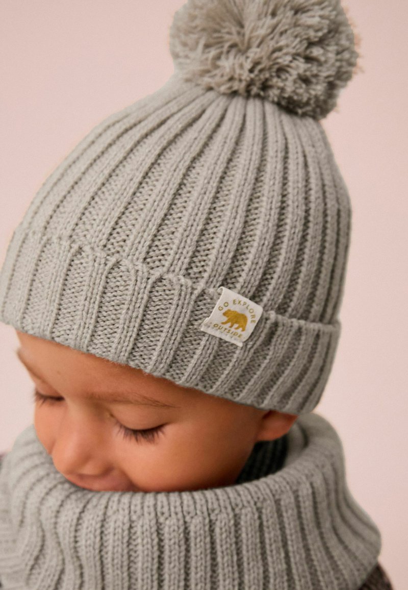 Lichtgrijze gebreide beanie met een ribbeltextuur en een pluizige pompon bovenop. Heeft een label met een beer en de tekst "GA BUITEN VERKENNEN."
