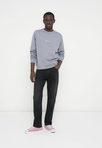 Ανοιχτό γκρι φούτερ με στρογγυλή λαιμόκοψη και λευκό κείμενο, συνδυασμένο με μαύρα τζιν και ροζ αθλητικά παπούτσια με λευκές σόλες και κορδόνια.