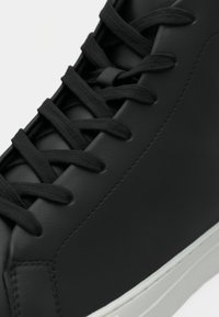 Sorte lædersneakers med flade sorte snøringer, glat tekstur og en hvid gummisål. Har et minimalistisk design og rund tåform.